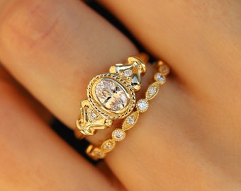 May include: A gold engagement ring with an oval-shaped diamond set in a delicate, ornate design. The ring is paired with a matching band featuring small diamonds.