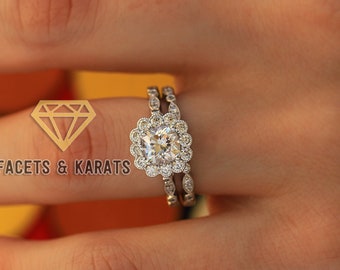 May include: A close-up of a diamond engagement ring set with a square-shaped center stone and a halo of smaller diamonds. The ring is set on a white gold band and is worn on a person's finger.