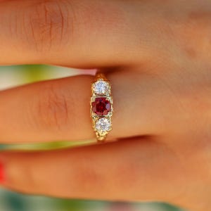 May include: A gold ring featuring a central round-cut red gemstone flanked by two smaller round-cut clear white gemstones. The metal band displays an intricate, textured pattern along the sides, holding the three stones in a linear arrangement.