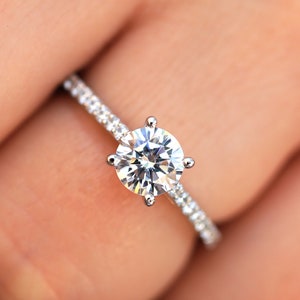 Puede incluir: Un anillo de compromiso de plata con un diamante central redondo y claro y una banda de diamantes más pequeños.