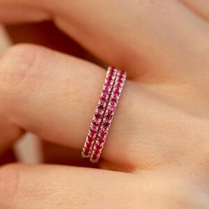 Puede incluir: Dos delicados anillos con una fila continua de pequeñas y vibrantes piedras preciosas rosas. Los anillos están engarzados en metal plateado o blanco, creando un aspecto brillante y elegante. Los anillos se muestran en el dedo de una persona.