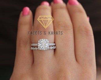 May include: A silver engagement ring set with a square-cut diamond centre stone surrounded by smaller diamonds. The ring is set on a band with a row of smaller diamonds. The ring is worn on a hand with pink nail polish.