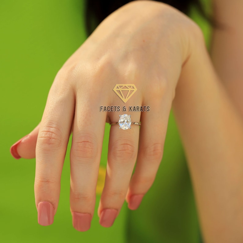 May include: A silver ring featuring an oval-cut diamond. The ring is worn on a hand with painted nails. The background is a solid green. The logo "FACETS & KARATS" is in the upper center.