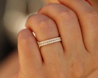 May include: Two gold-colored eternity rings, each encrusted with small, clear gemstones. The rings are stacked on a hand with red nail polish. The rings are in focus, with a blurred background.