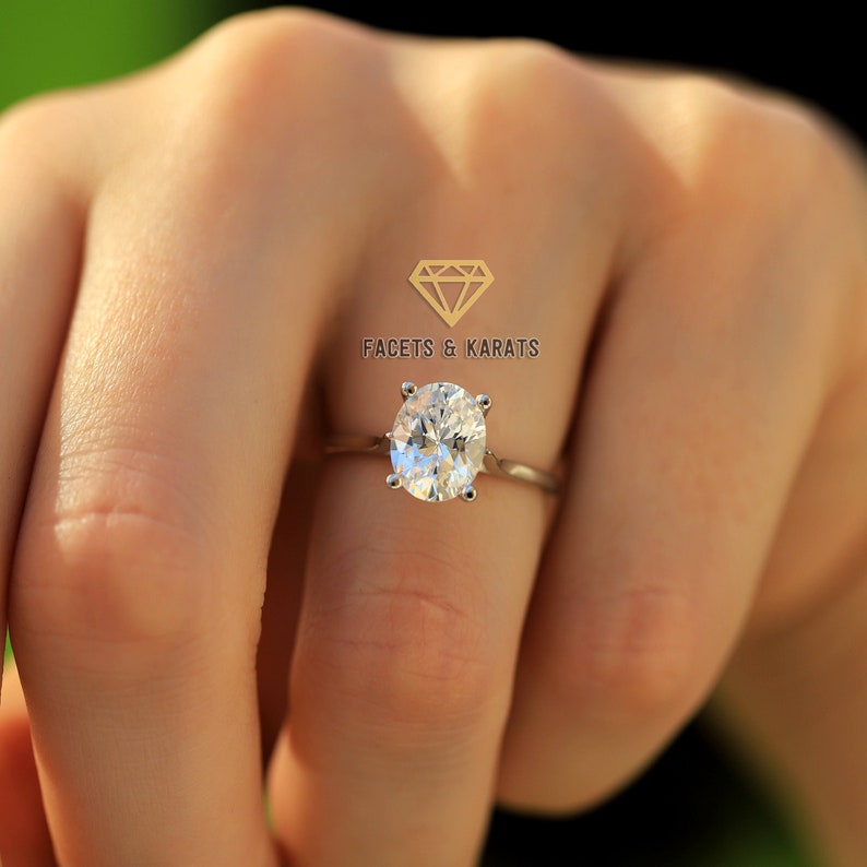 May include: A silver ring featuring an oval-cut diamond. The ring is set on a thin band and is being worn on a finger. The logo "FACETS & KARATS" is visible in the upper right corner.