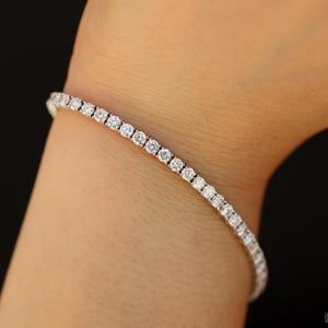 a 14k white gold diamond tennis bracelet is dangling on the wrist of a model. The stones are round cut diamonds of 3mm diameter. The setting style is 4-prong with double basket wire setting. You can see the Facets and Karats logo.