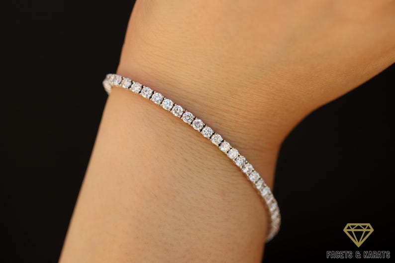 a 14k white gold diamond tennis bracelet is dangling on the wrist of a model. The stones are round cut diamonds of 3mm diameter. The setting style is 4-prong with double basket wire setting. You can see the Facets and Karats logo.
