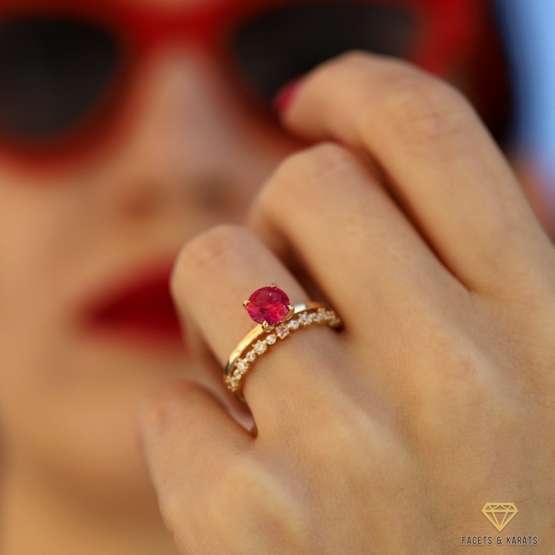 May include: A gold ring with a large, round, red gemstone surrounded by smaller, white gemstones. The ring is worn on the finger of a person wearing red lipstick.