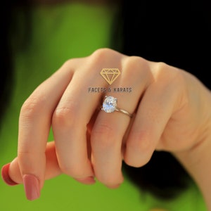 May include: A silver ring featuring an oval-cut diamond. The ring is displayed on a slender band. The image includes the text "FACETS & KARATS" and a diamond logo. The background is blurred with green and black.