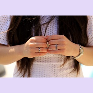 May include: A woman's hands wearing several rings, including a diamond ring and a band ring. The rings are on her left hand. She is wearing a white dress with a lace pattern.