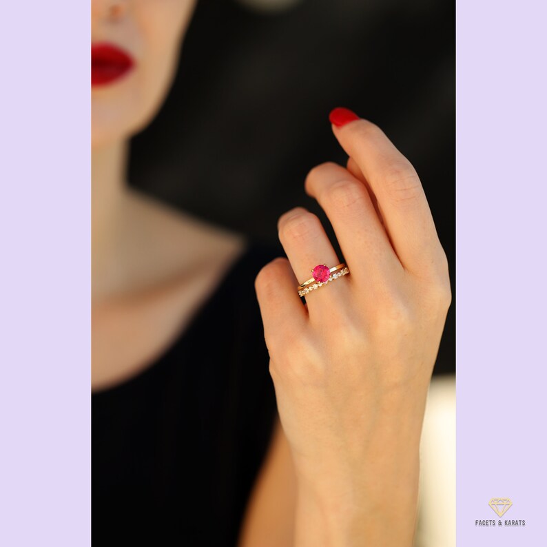 May include: A gold ring featuring a single, round, red gemstone encircled by smaller, clear gemstones. The ring is worn on the ring finger of a hand with red nail polish.