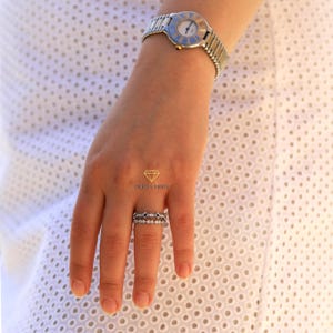 May include: A close-up of a hand wearing two silver rings with small diamonds. The rings are on the ring finger of the left hand. The hand is wearing a silver watch with a blue face.
