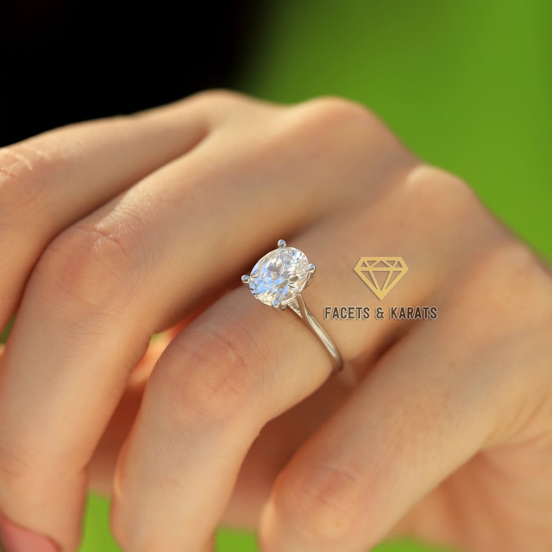 May include: A silver engagement ring featuring an oval-cut diamond, held in a hand. The ring's design is simple, highlighting the brilliance of the gemstone. The image includes the text "FACETS & KARATS".