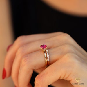 May include: A gold ring set with a single red gemstone and a band of smaller diamonds. The ring is worn on a person's finger.