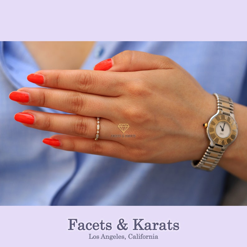 Op de afbeelding: Een delicate gouden ring met een rij kleine diamanten. De ring zit aan een vinger met rode nagellak.