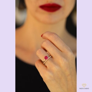 May include: A gold ring with a red gemstone and a smaller band of diamonds. The ring is on a person's finger.