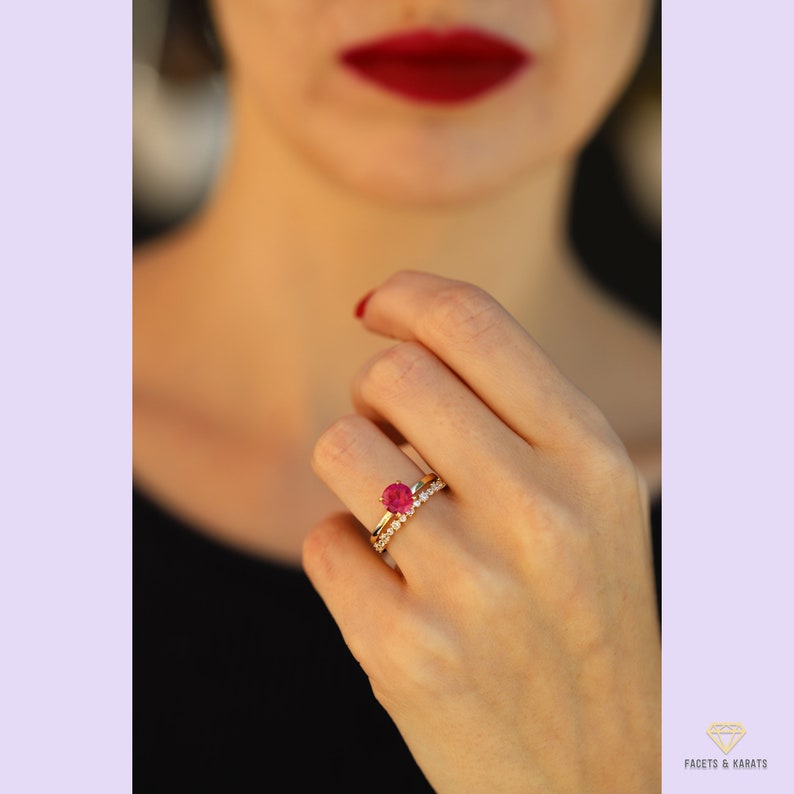 May include: A gold ring with a red gemstone and a smaller band of diamonds. The ring is on a person's finger.