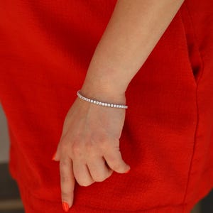 May include: A silver tennis bracelet with small, clear cubic zirconia stones. The bracelet is on a woman's wrist.