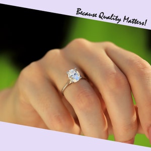 May include: A silver engagement ring featuring an oval-cut diamond. The ring is worn on a finger, with the text "Because Quality Matters!" visible. The background is a blurred green, suggesting an outdoor setting.