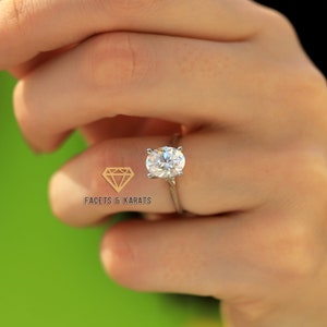 May include: A silver ring featuring a large, oval-cut diamond. The ring has a slender band. The image includes the logo "FACETS & KARATS" in the lower left corner. The background is a soft green.
