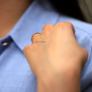 Op de afbeelding: Een gouden bandring met een textuurontwerp. De ring wordt gedragen aan de vinger van een persoon die een blauw shirt draagt. De tekst "FACETS & KARATS" is zichtbaar op de ring.