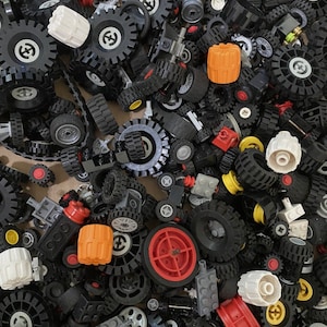 May include: A large collection of black Lego wheels and tires of various sizes and styles. Some of the wheels have red, orange, and yellow accents. The wheels are scattered on a brown surface.