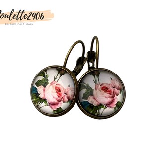 May include: A pair of round earrings with a floral design. Each earring features a pink rose print set against a white background, framed by a bronze-coloured metal. The earrings have a lever-back closure. The text "Poulette2906 BIJOUX FAIT MAIN" is in the upper left corner.