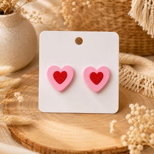 May include: A pair of stud earrings attached to a white display card. Each earring features a layered heart design with a light pink outer heart and a smaller, textured red glitter inner heart, creating a distinct two-tone appearance.
