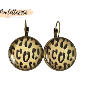 Puede incluir: Un par de pendientes redondos con estampado de leopardo, con marcos de metal bronce y cierres de palanca. Los pendientes tienen un fondo beige con manchas marrones oscuras. El texto "Poulette2906 BIJOUX FAIT MAIN" es visible.