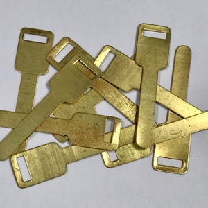 May include: A pile of small, rectangular brass tags, each with a keyhole-shaped aperture at the top. The tags have a slightly worn look, possibly vintage or used. They are scattered on a white surface.