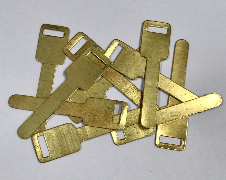 May include: A pile of small, rectangular brass tags, each with a keyhole-shaped aperture at the top. The tags have a slightly worn look, possibly vintage or used. They are scattered on a white surface.