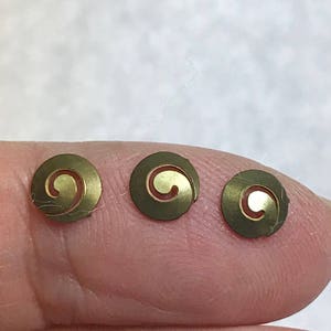 May include: Three small, circular brass embellishments with a spiral design. Each piece is a flat disc with a raised, swirling interior. The brass has a slightly aged appearance, and the spiral is a lighter gold color.