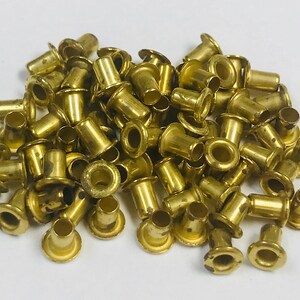 May include: A pile of small, brass-colored grommets. Each grommet has a flared base and a cylindrical barrel, with a hole in the center. The grommets are scattered on a white surface.