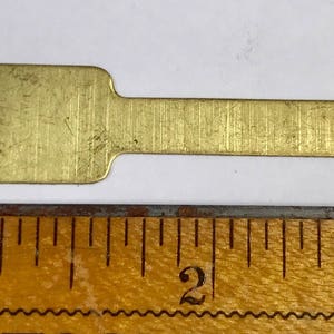 May include: A small, rectangular brass key blank with a rectangular hole and a rounded end. The key blank is approximately 5 cm long and lies next to a wooden ruler.