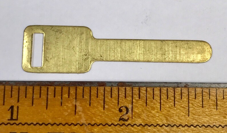May include: A small, rectangular brass key blank with a rectangular hole and a rounded end. The key blank is approximately 5 cm long and lies next to a wooden ruler.