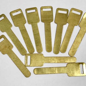 May include: A collection of ten brass-coloured key blanks arranged on a white surface. Each key blank has a rectangular head with a rectangular hole and a long, flat body. The keys are of similar size and shape.
