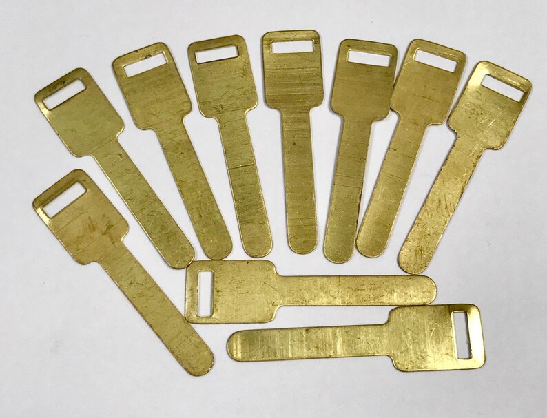 May include: A collection of ten brass-coloured key blanks arranged on a white surface. Each key blank has a rectangular head with a rectangular hole and a long, flat body. The keys are of similar size and shape.
