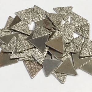 May include: A collection of small, metallic triangle-shaped objects. Some triangles have a textured surface, while others are smooth. The objects are scattered on a white surface, creating a random pattern. The color is a muted silver.