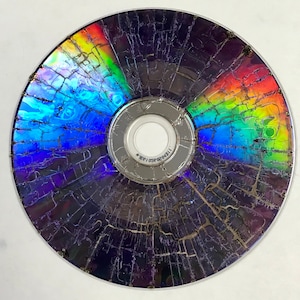 May include: A cracked CD with a rainbow-coloured, iridescent surface. The disc's colours are visible through the cracks, with a silver centre and a white inner circle. The disc's surface is heavily damaged.