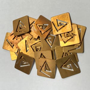 Peut inclure: Un ensemble de petites découpes carrées en métal de couleur laiton. Chaque découpe présente un motif triangulaire avec une spirale à l'intérieur. Les découpes sont dispersées sur une surface claire, avec des nuances d'or et de bronze.