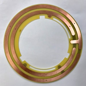 May include: A circular electronic component with a pale yellow base and copper traces. The component is ring-shaped with an open centre and cutouts. The copper traces are arranged in concentric arcs around the centre.
