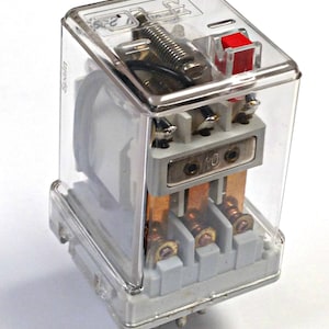 May include: A clear, rectangular electrical relay with a transparent cover. It features copper contacts, a red button, and a silver spring. The relay is mounted on a gray base with pins.