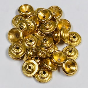 Könnte beinhalten: Eine Ansammlung kleiner, goldfarbener Metallperlenkappen. Jede Kappe hat eine ausgestellte, konische Form mit einem zentralen Loch und einem dekorativen, kreisförmigen Design auf der flachen Oberseite. Die Kappen sind auf einer weißen Oberfläche verstreut.