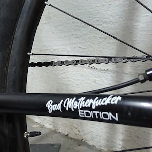 Puede incluir: Primer plano de una rueda de bicicleta negra con una cadena plateada y una pegatina negra que dice "Bad Motherfucker Edition".