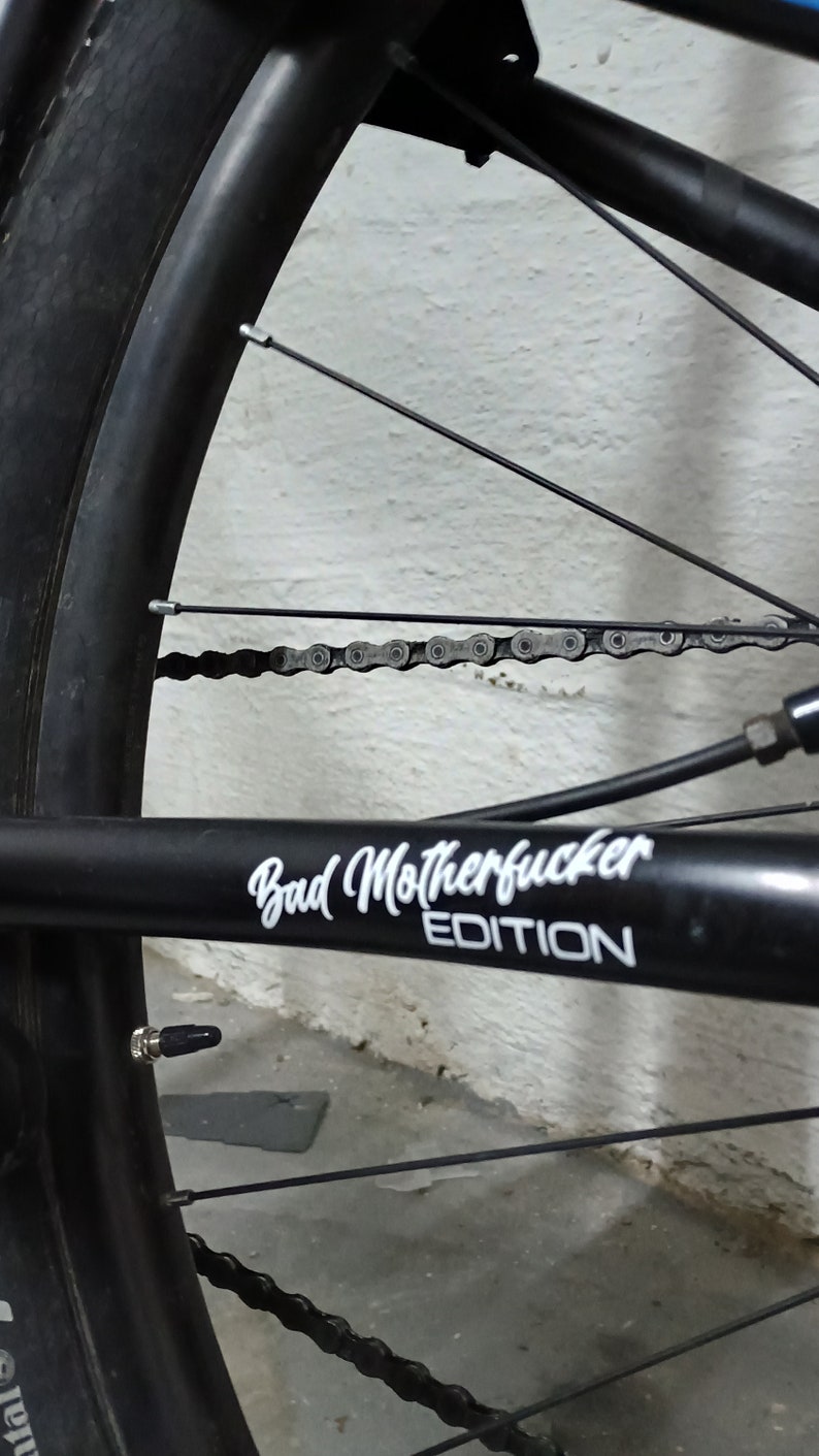 Puede incluir: Primer plano de una rueda de bicicleta negra con una cadena plateada y una pegatina negra que dice "Bad Motherfucker Edition".