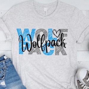 May include: A light gray t-shirt with the word "WOLF PACK" in blue, black, and gray, with a heart symbol. The shirt is paired with denim shorts and gray sneakers. A small potted plant is also visible.