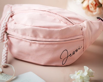 May include: A pink fanny pack with a white drawstring and a black zipper. The words "Jessi" are embroidered on the front of the bag.