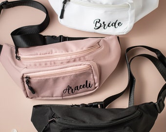 May include: Three fanny packs with adjustable straps. The white fanny pack says "Bride" in black lettering. The pink fanny pack says "Araceli" in black lettering. The black fanny pack says "Jessica" in white lettering.