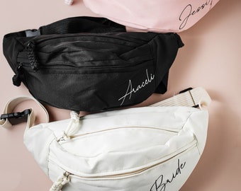 May include: Three fanny packs in different colors: pink, black, and white. The pink fanny pack has the name "Jessi" embroidered on it. The black fanny pack has the name "Araceli" embroidered on it. The white fanny pack has the word "Bride" embroidered on it.