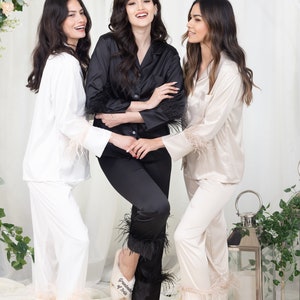 May include: Three women wearing matching satin pajamas with feather trim. The pajamas are white, black, and beige. The women are wearing matching white slippers with embroidered text. The text on the slippers reads "bridesmaid", "ruschkin", and "their".